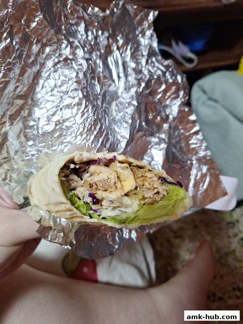 A person holds a foil-wrapped wrap filled with chicken, lettuce, and cabbage.