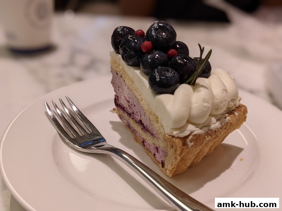 A slice of blueberry cake with whipped cream and fresh blueberries on a white plate with a fork.