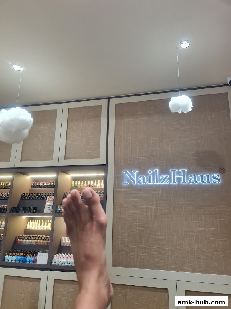 A foot is positioned in front of a wall with the NailzHaus logo and shelves of nail polish.