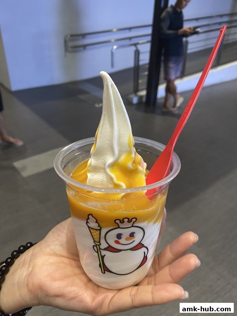 A hand holds a cup of soft-serve ice cream with mango topping and a red spoon.