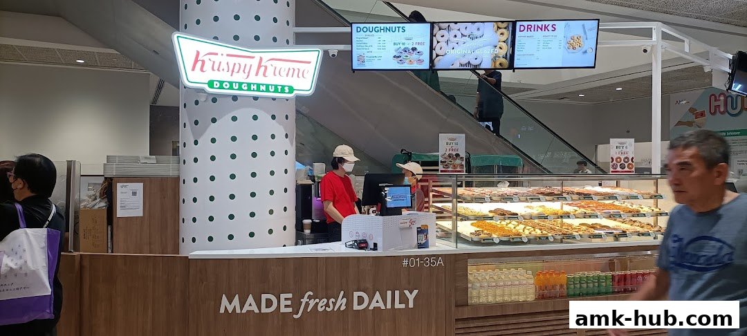 Krispy Kreme doughnut shop with a display of fresh doughnuts and drinks, and staff serving customers.