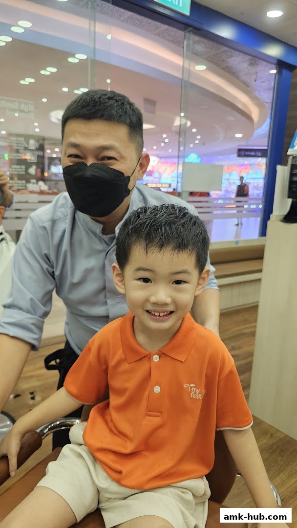 A smiling young boy in an orange shirt sits in a barber chair with a masked barber behind him.