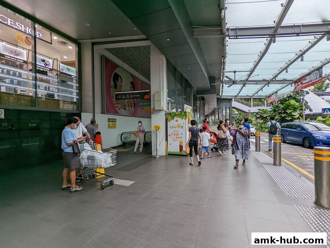 People walking along a covered walkway outside shops, with a blue car parked nearby.