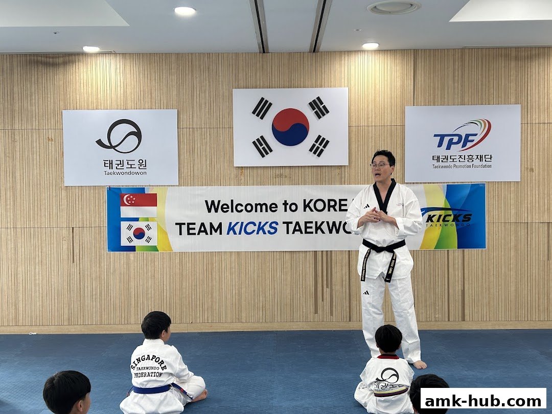 A Taekwondo instructor addresses young students in a training room with national flags and logos on the wall.