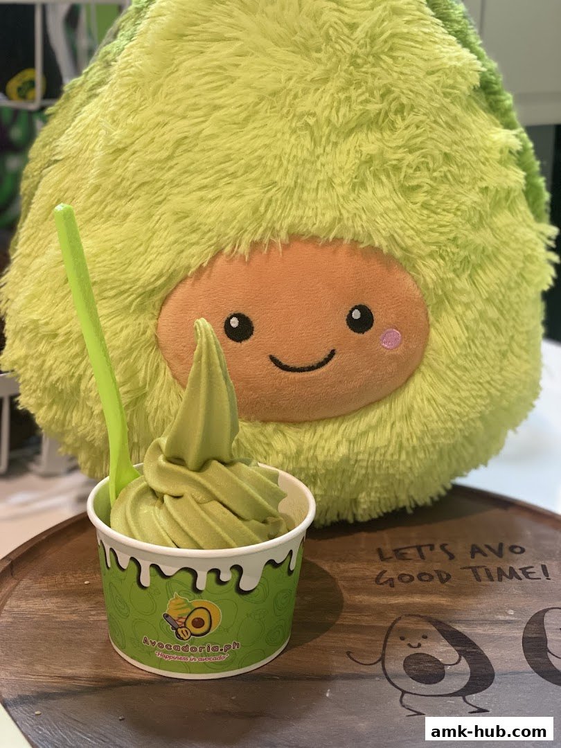 A cup of green soft serve ice cream with a cute avocado plush toy in the background.