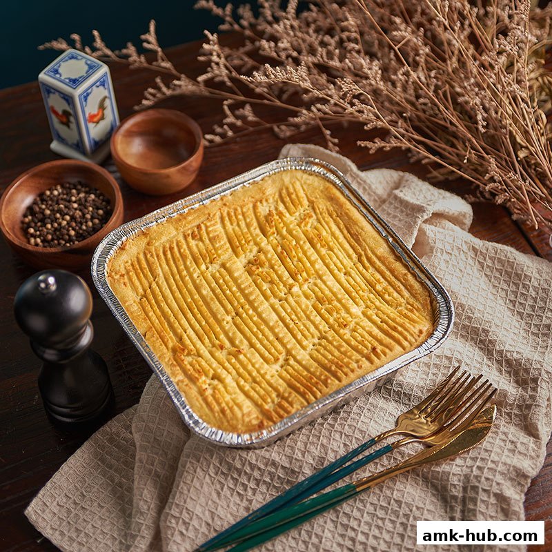 A golden-brown shepherd's pie in a foil container, ready to serve, with cutlery and spices.