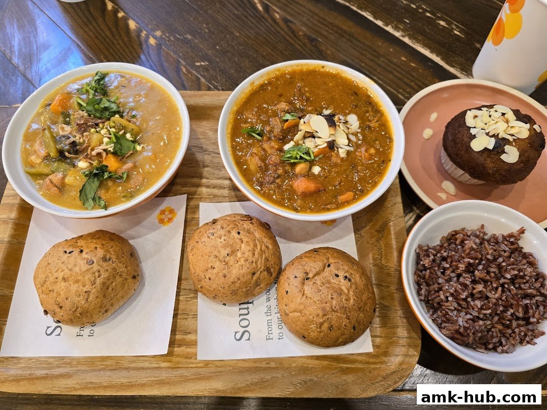 A wooden tray holds two bowls of soup, three bread rolls, a muffin, and a bowl of rice.