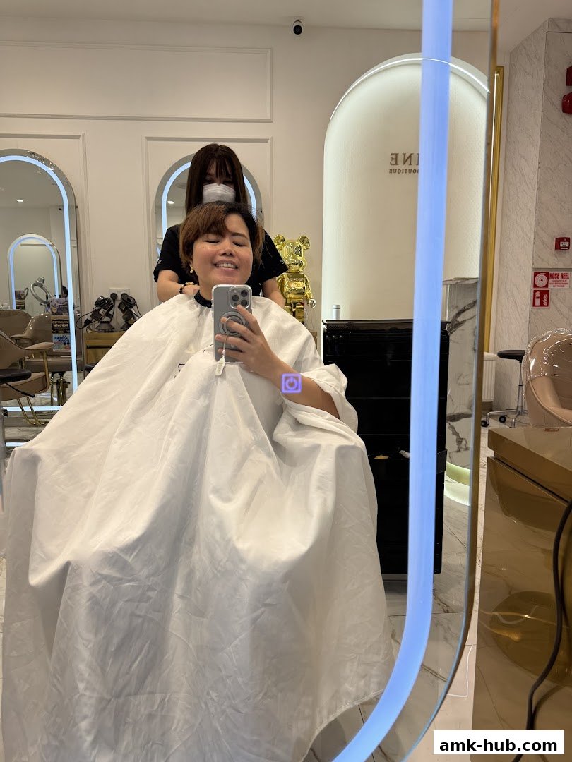 A person smiles while taking a selfie in a salon chair, with a stylist behind them.