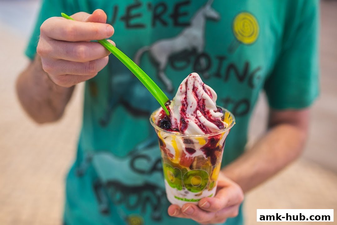 A person holds a cup of frozen yogurt with fruit and sauce, being eaten with a green spoon.