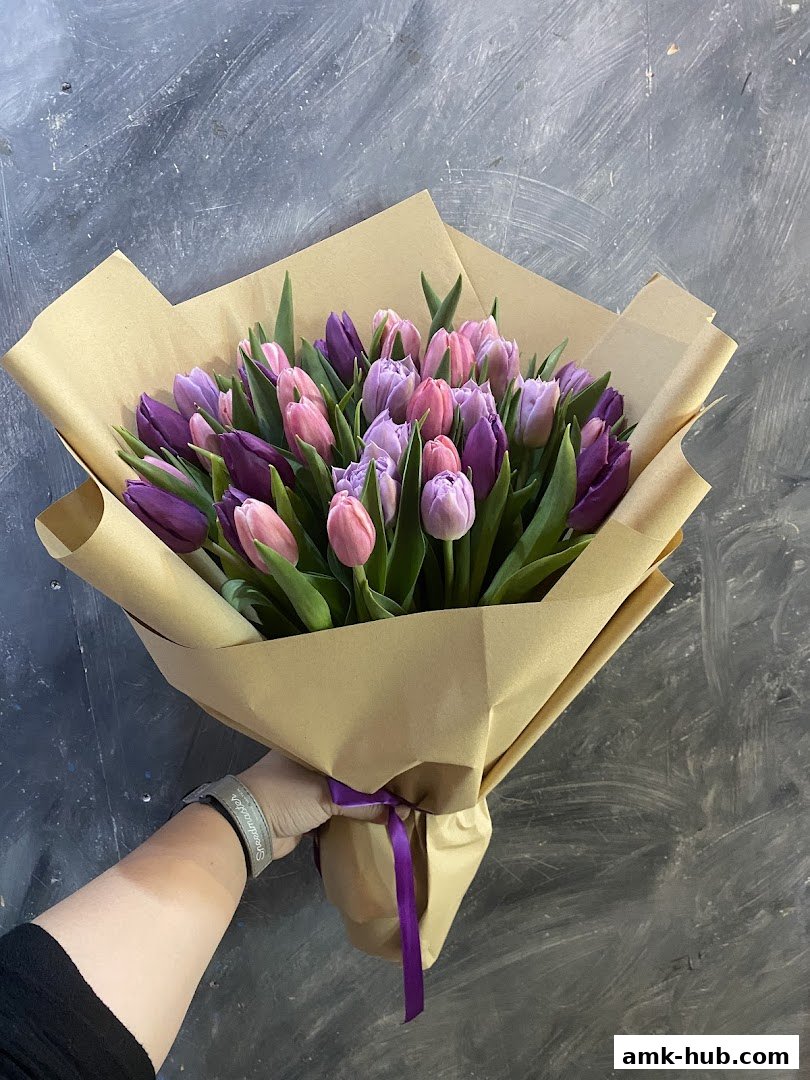 A hand holding a beautiful bouquet of purple and pink tulips wrapped in brown paper.