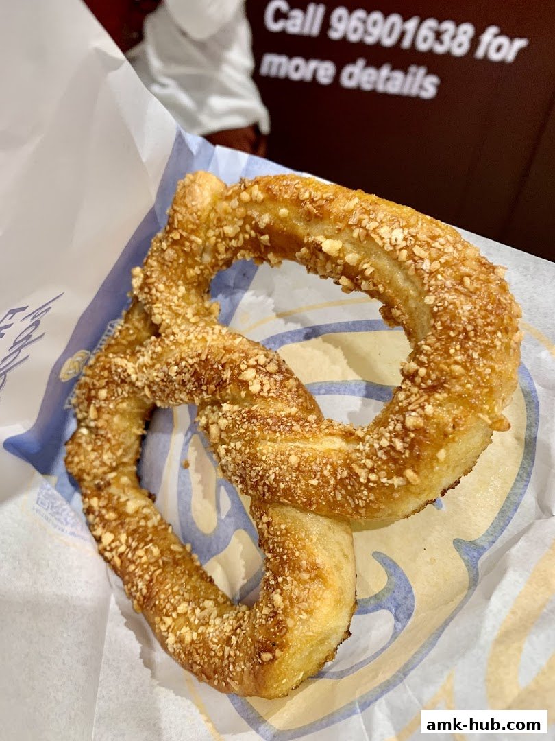 A close-up of a pretzel covered in nuts, served in branded paper.