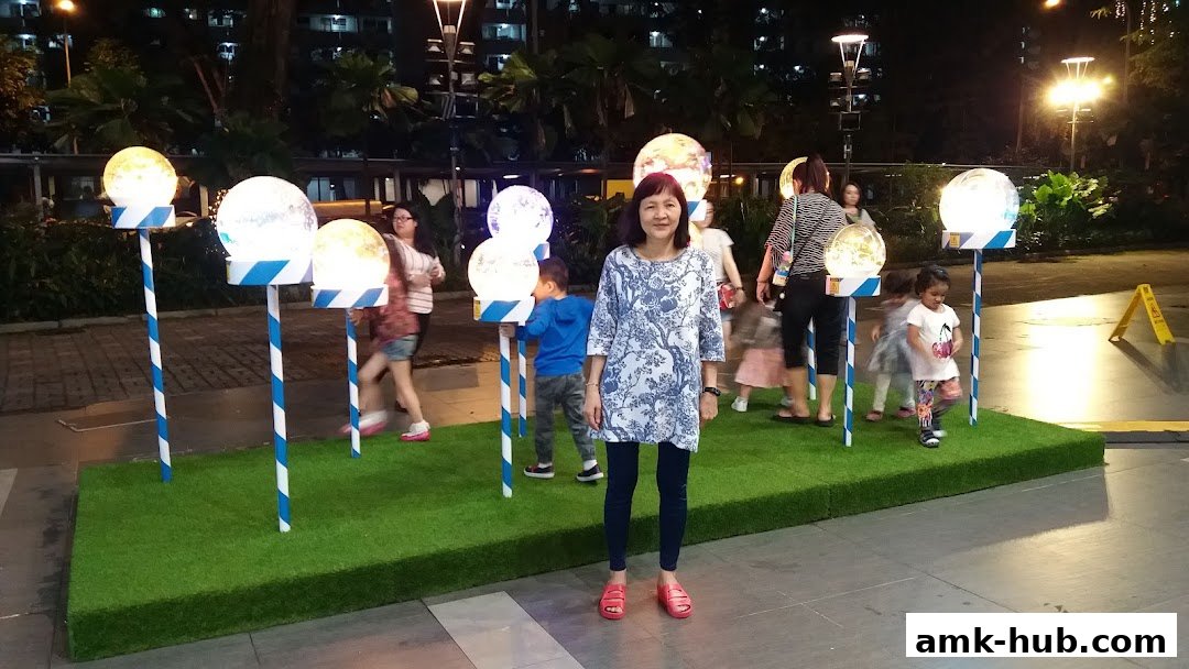 A woman stands in front of decorative glowing orbs on poles, with people walking in the background.