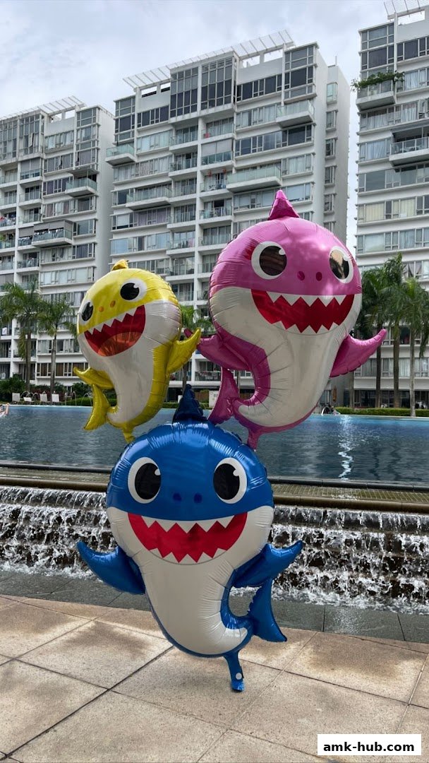 Three Baby Shark balloons float in front of a swimming pool and modern buildings.