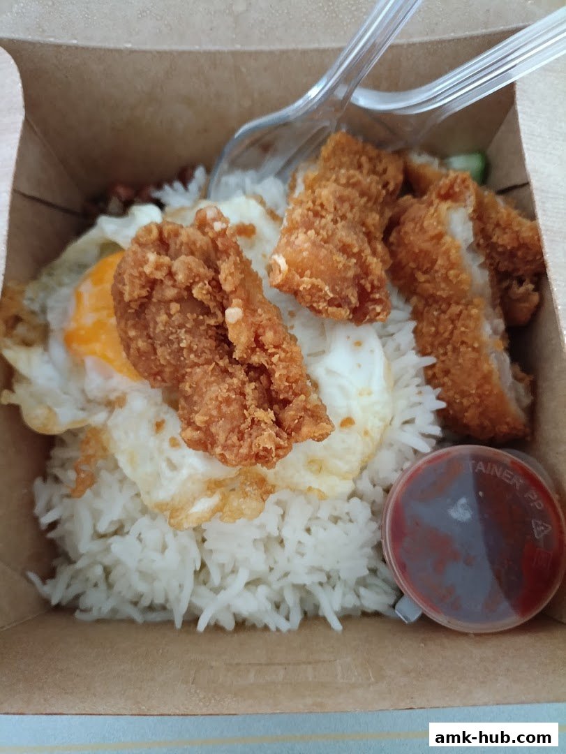 A close-up of a Nasi Lemak dish with fried chicken, rice, and a fried egg in a takeaway box.