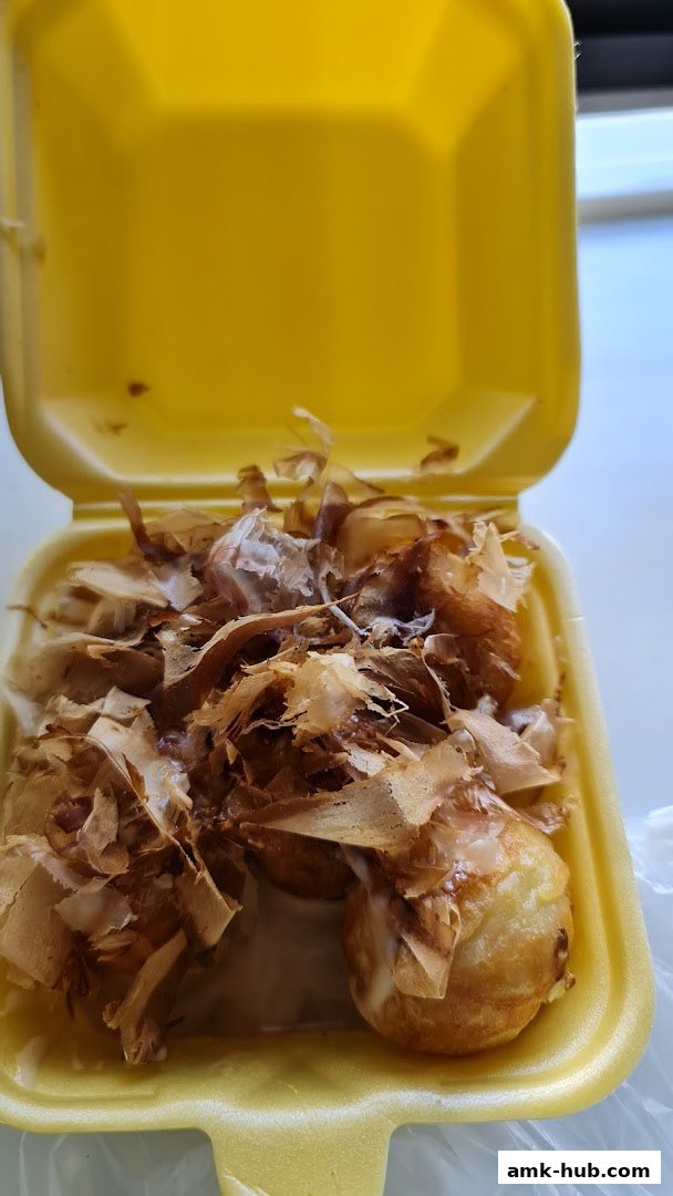 A close-up of takoyaki balls topped with bonito flakes in a yellow takeout container.