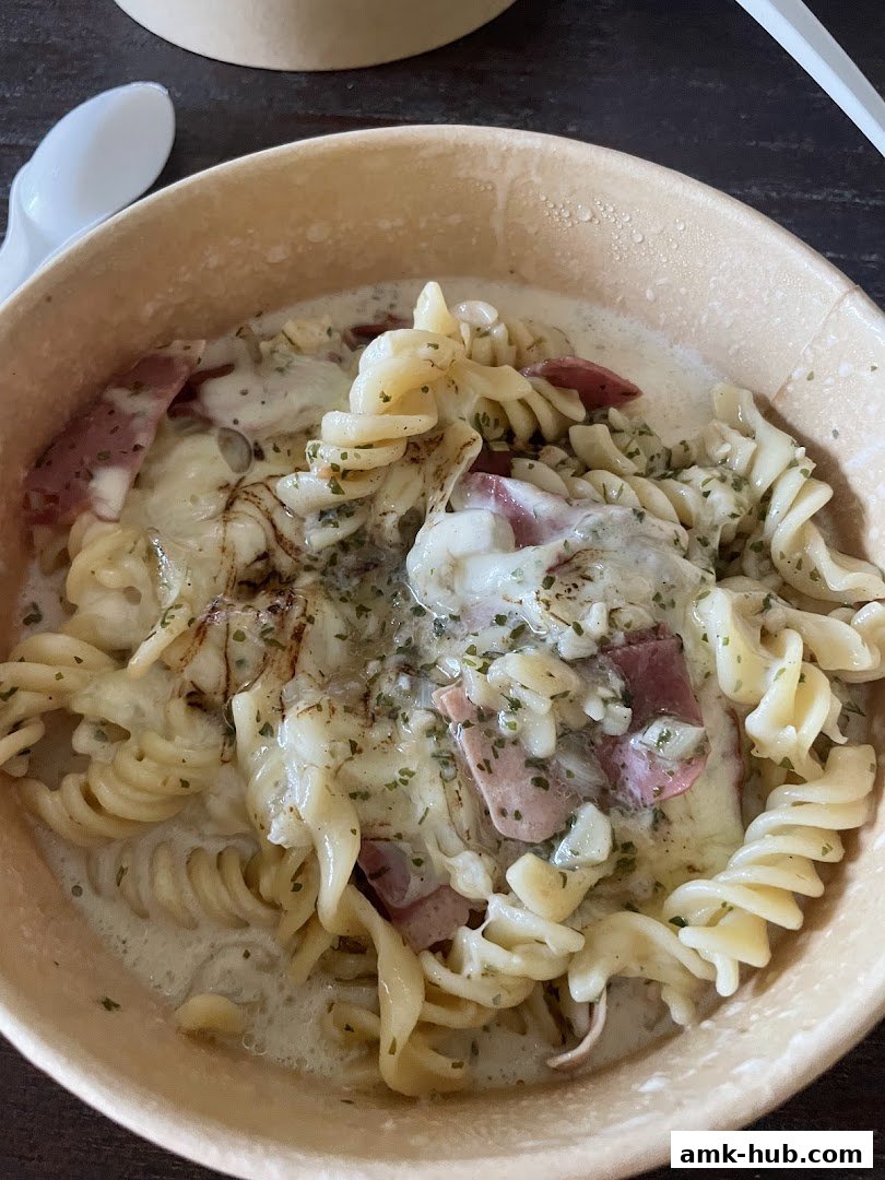 A close-up shot of a creamy pasta dish with ham and cheese in a disposable container.