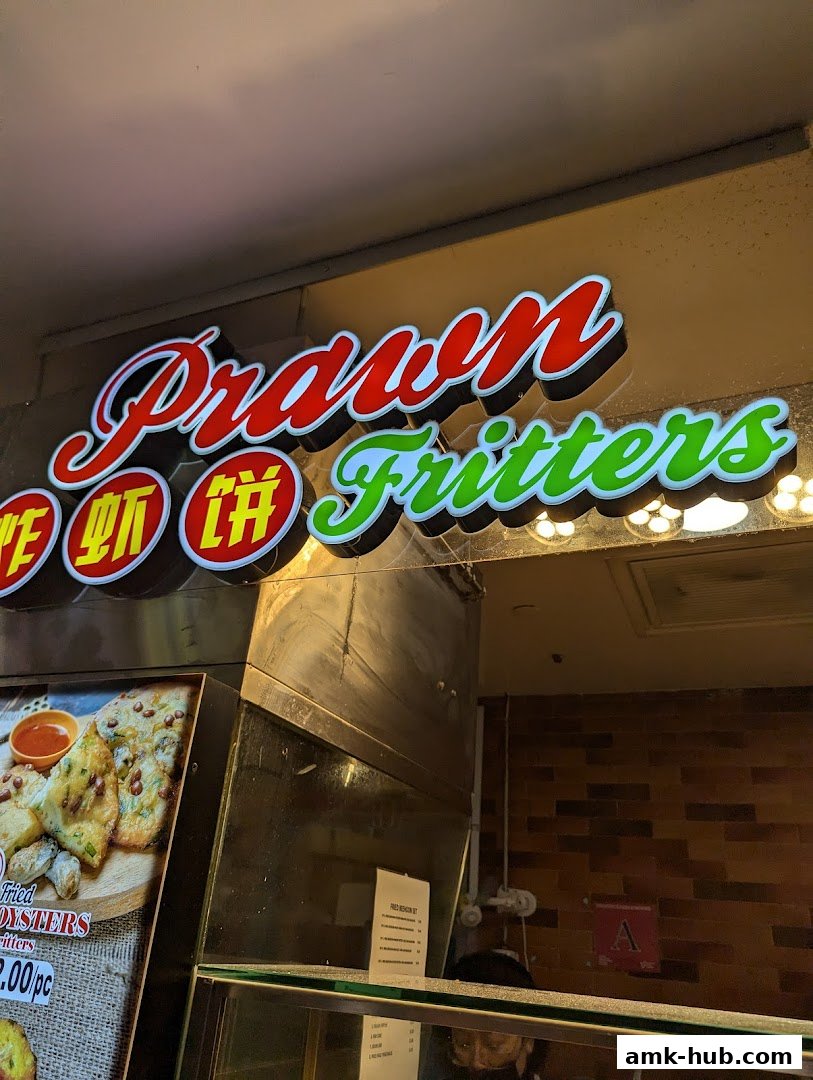 A brightly lit sign for 'Prawn Fritters' above a food stall with a display of food.