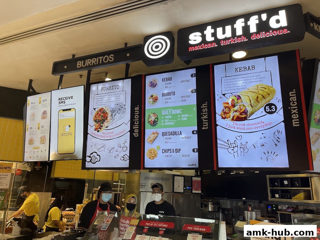 A food stall with digital menus displaying burritos, kebabs, and daily bowls, with staff serving customers.