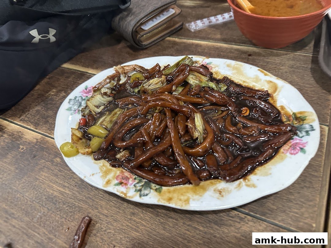 A plate of Hokkien Mee with vegetables and a bowl of soup on a wooden table.