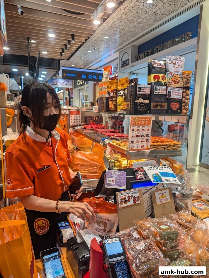 A shop assistant in an orange uniform stands behind a counter filled with various packaged snacks and pastries.