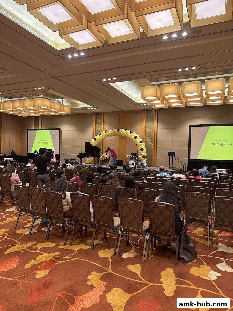 An audience watches a performance on stage with a piano, drums, and balloon arch.