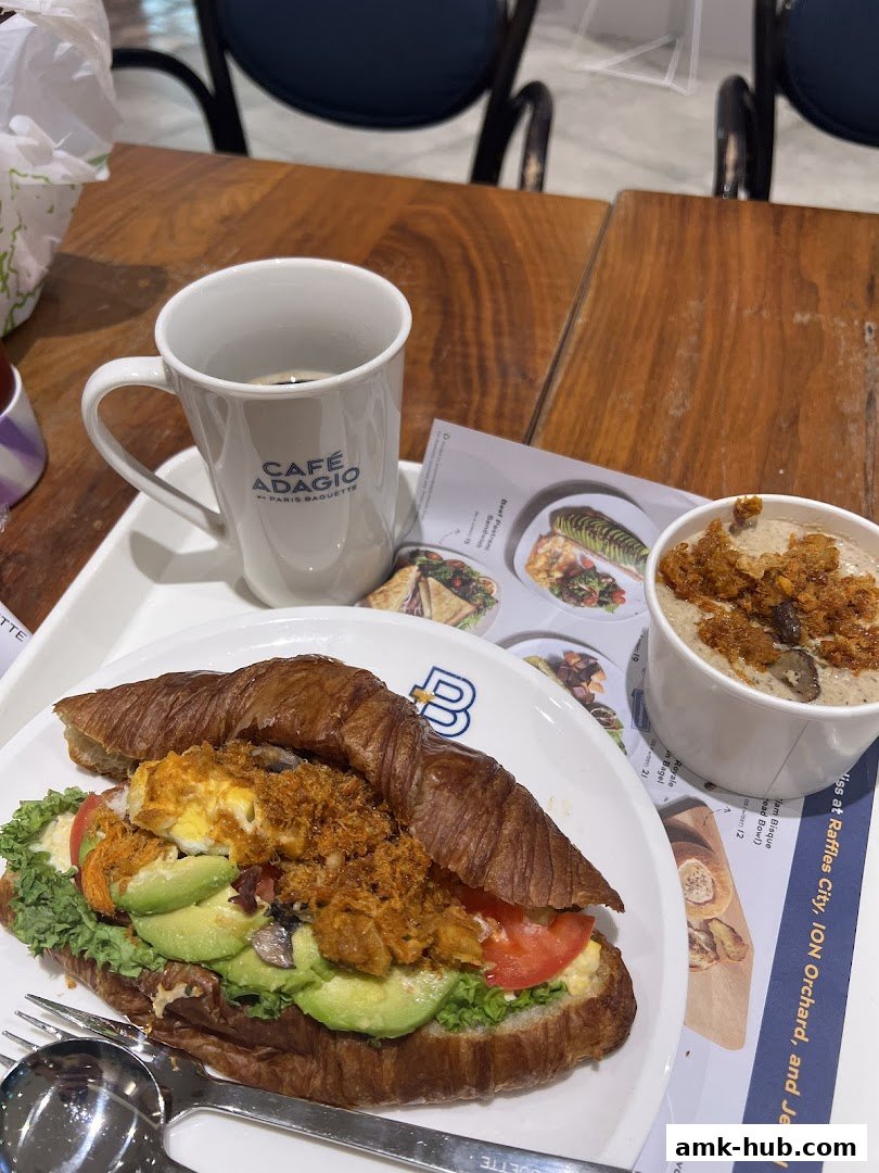 A croissant sandwich with avocado, tomato, and shredded meat, served with coffee and soup.