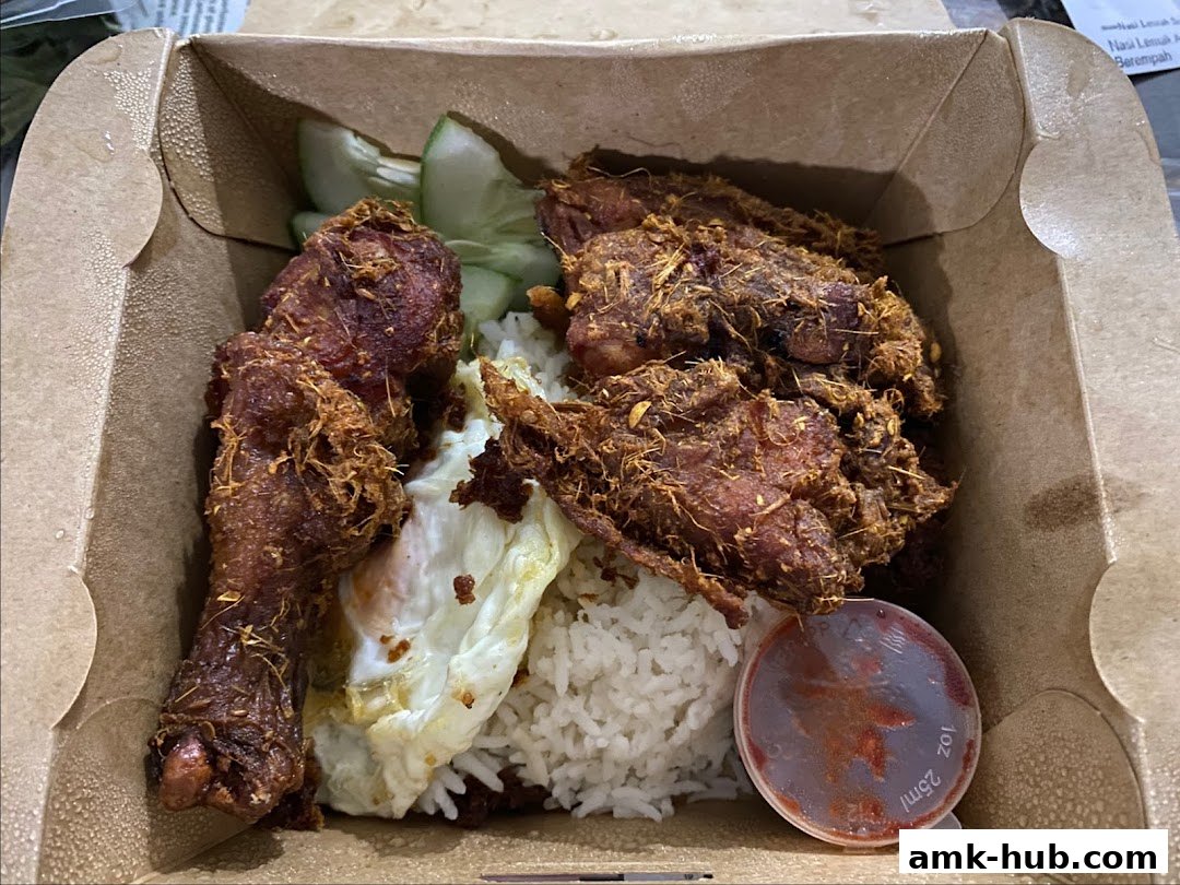 A takeaway box filled with Nasi Lemak, featuring fried chicken, rice, egg, cucumber, and chili sauce.