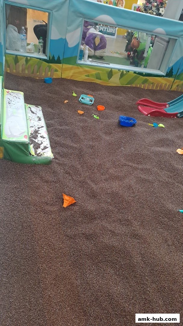 An indoor play area with sand, toys, and a slide, featuring cartoon characters visible through windows.