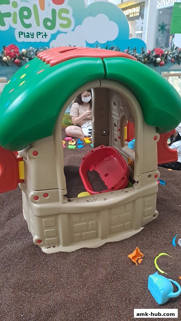 A child's playhouse structure in a sandpit with toys and a person wearing a mask.