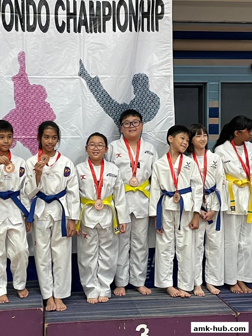 Children in Taekwondo uniforms with medals stand on a podium under a championship banner.