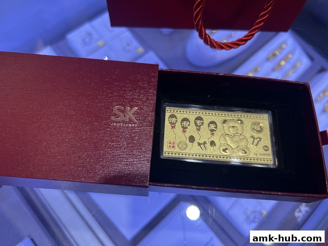 A gold bar with a teddy bear design is presented in a red gift box from SK Jewellery.