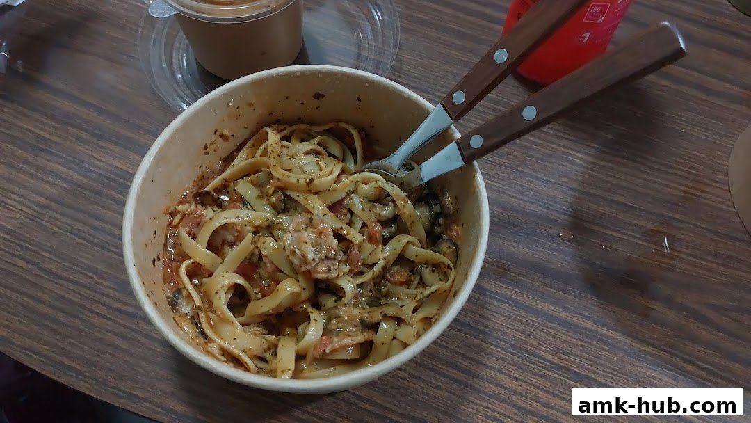 A bowl of fettuccine pasta with sauce and ingredients, served with cutlery.