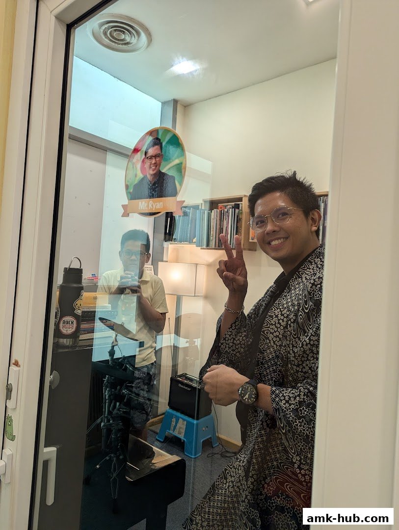 Two men are in a room with musical instruments and bookshelves, one smiling at the camera.