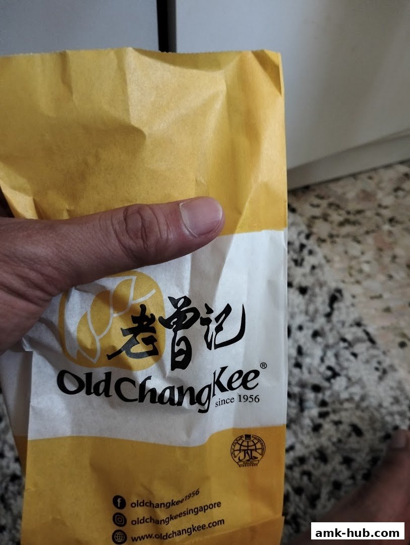 A hand holds a yellow and white paper bag with the Old Chang Kee logo and website.