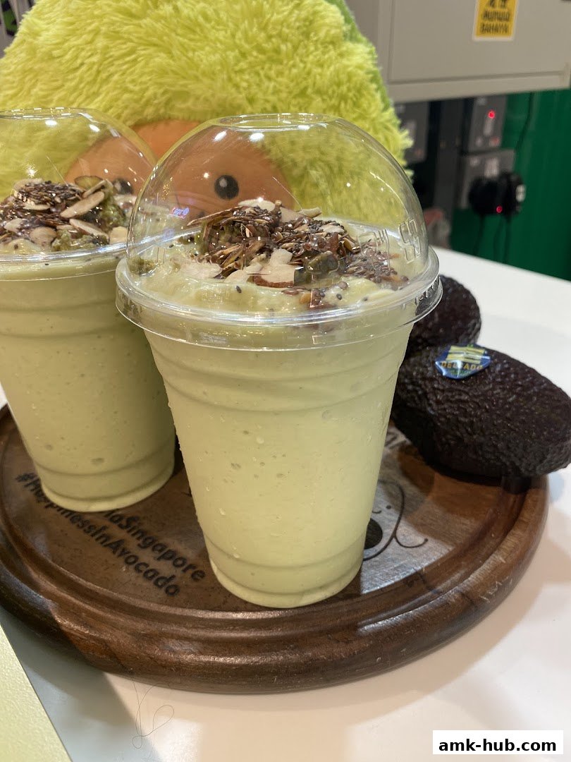 Two avocado smoothies topped with seeds and nuts on a wooden tray with avocados.