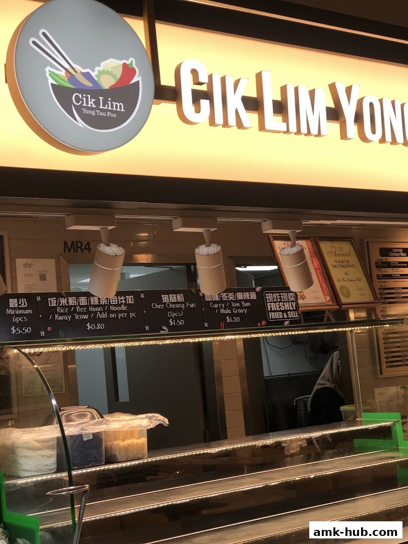 The front of a food stall named Cik Lim Yong Tau Foo with a bright illuminated sign and menu items displayed.