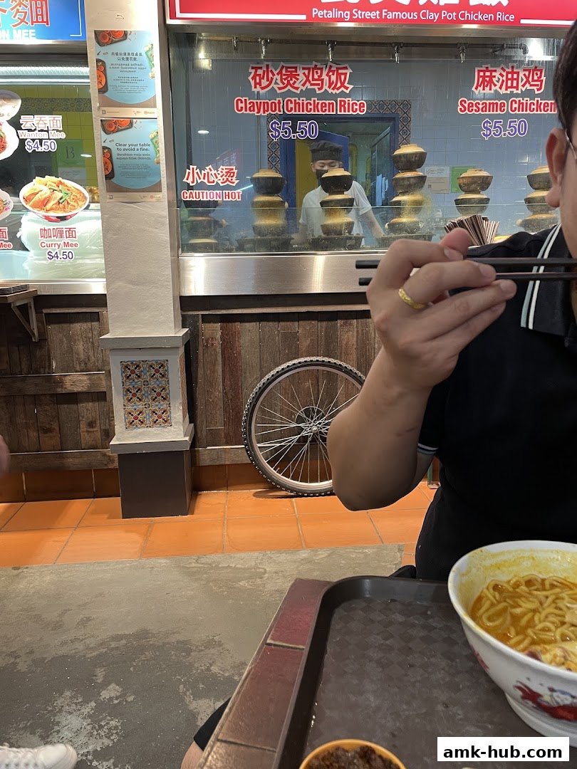 A person eating at a Malaysian food stall with various noodle dishes and claypot rice on display.