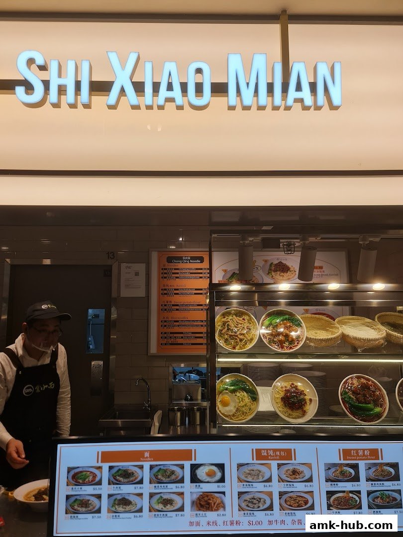 A food stall with a sign reading 'SHI XIAO MIAN' and a display of noodle dishes.