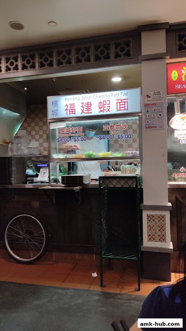 A food stall counter with a sign displaying menu items and prices, and a staff member preparing food.
