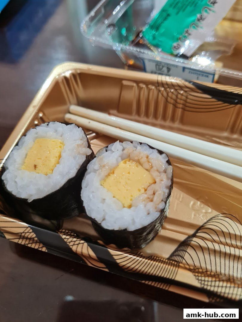 Two tamagoyaki sushi rolls in a takeaway container with chopsticks.