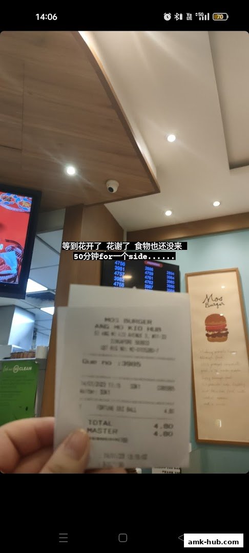 A receipt from MOS Burger is held up, with a digital display and a framed advertisement in the background.