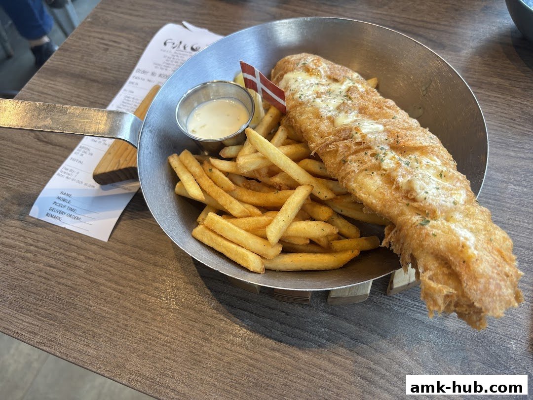 A serving of fish and chips with a side of sauce, presented in a metal pan.