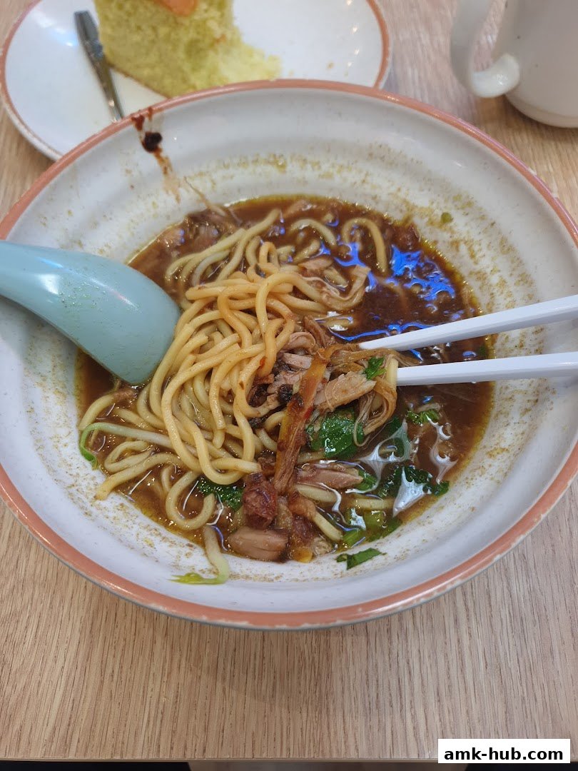 A close-up of a bowl of noodles with meat and greens, with a slice of cake in the background.