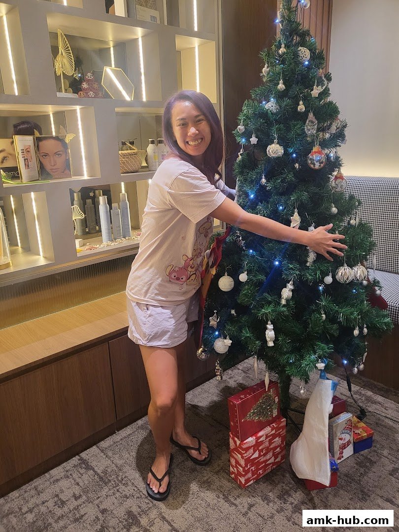 A woman poses happily next to a decorated Christmas tree with gifts underneath.