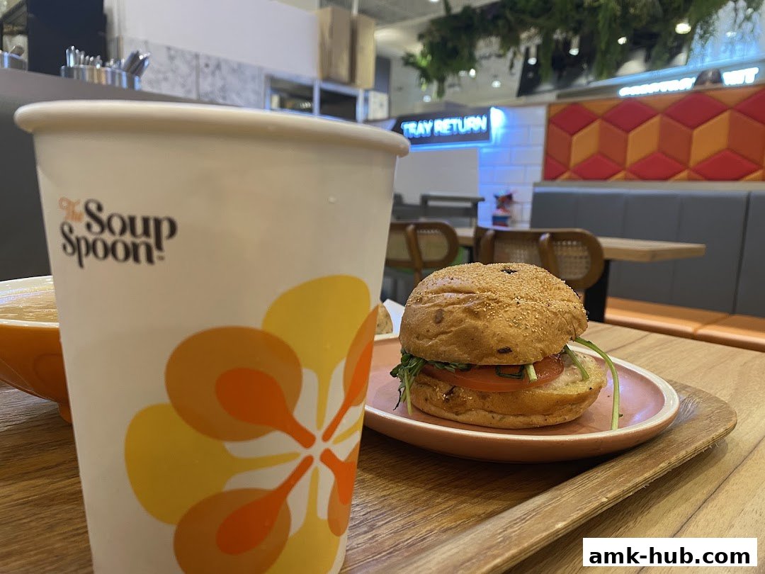 A close-up of a burger and a cup with the Soup Spoon logo on a wooden tray.