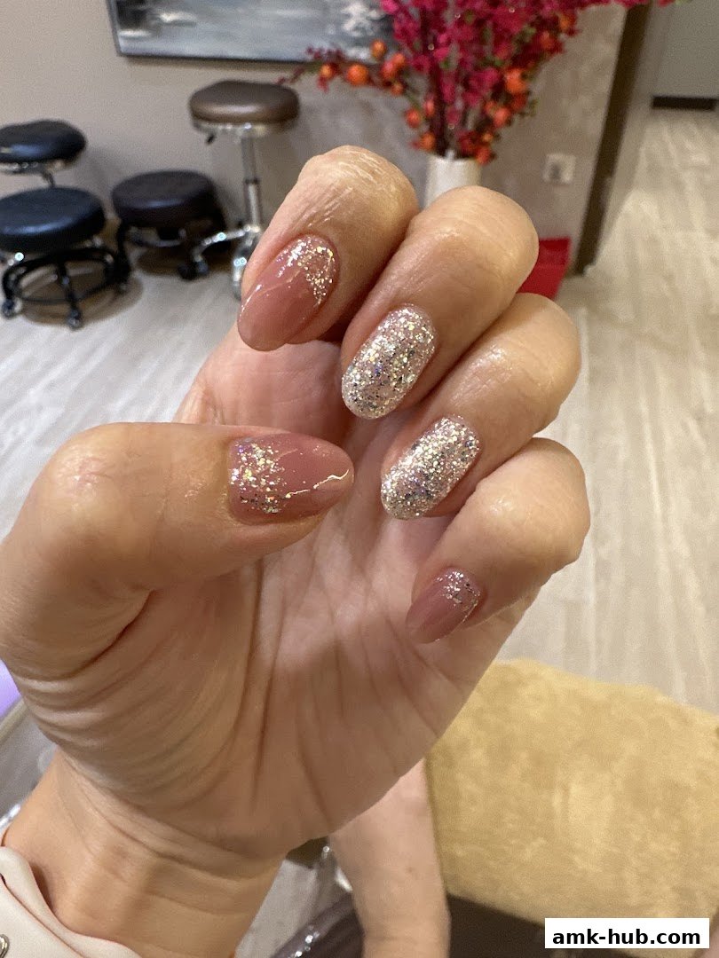 A close-up of a hand with beautifully manicured nails featuring a nude polish and glitter accents.