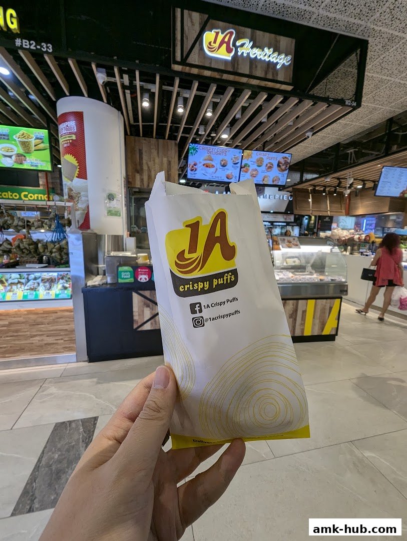 A hand holds a paper bag with the '1A Heritage crispy puffs' logo in front of the shop.