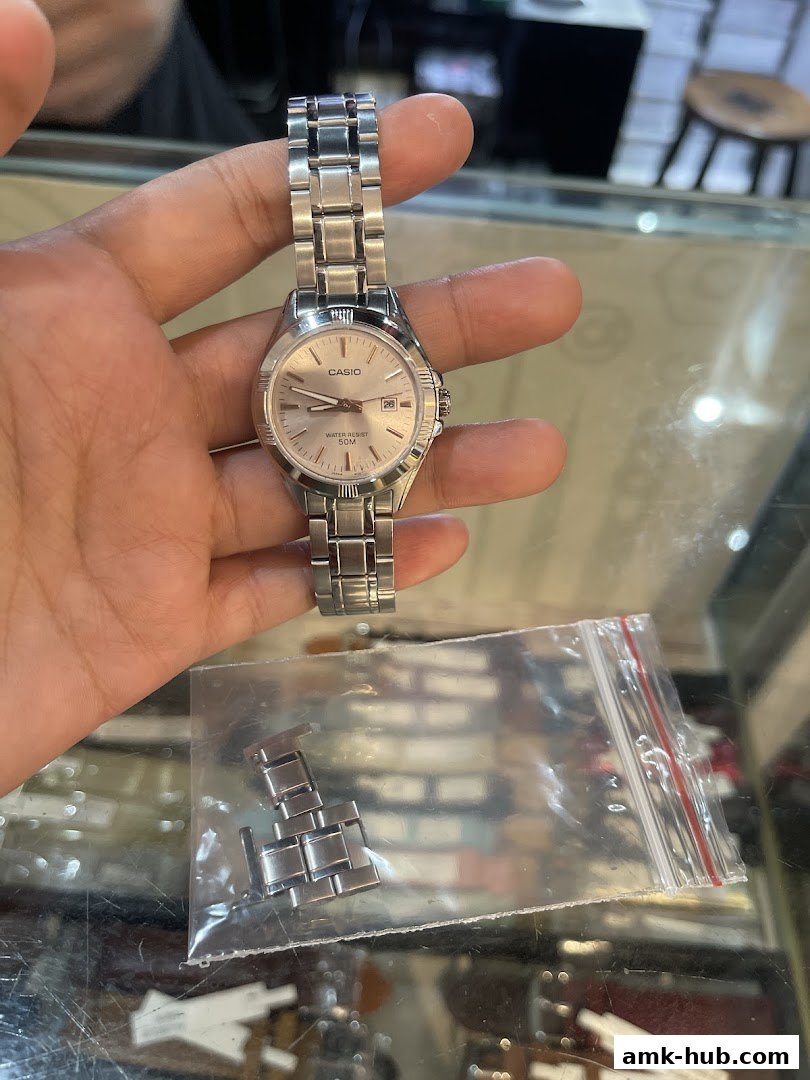 A hand holds a silver Casio watch with a metal band and extra links in a plastic bag.
