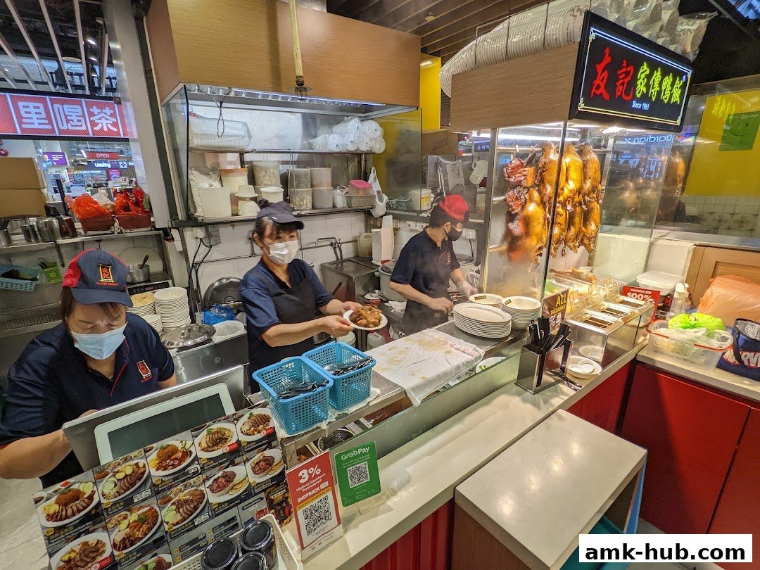 Staff preparing food at a roast duck stall with hanging ducks and a display of dishes.