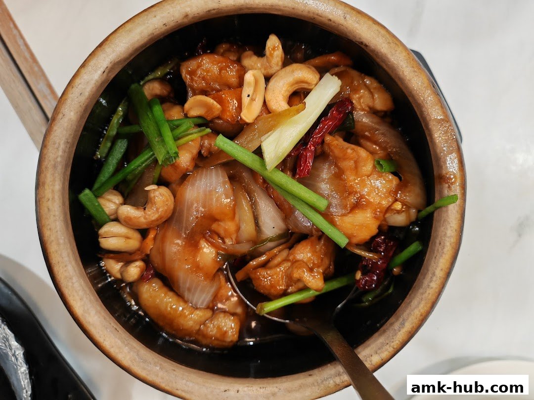 A close-up overhead view of a clay pot dish with chicken, cashews, onions, and chili.